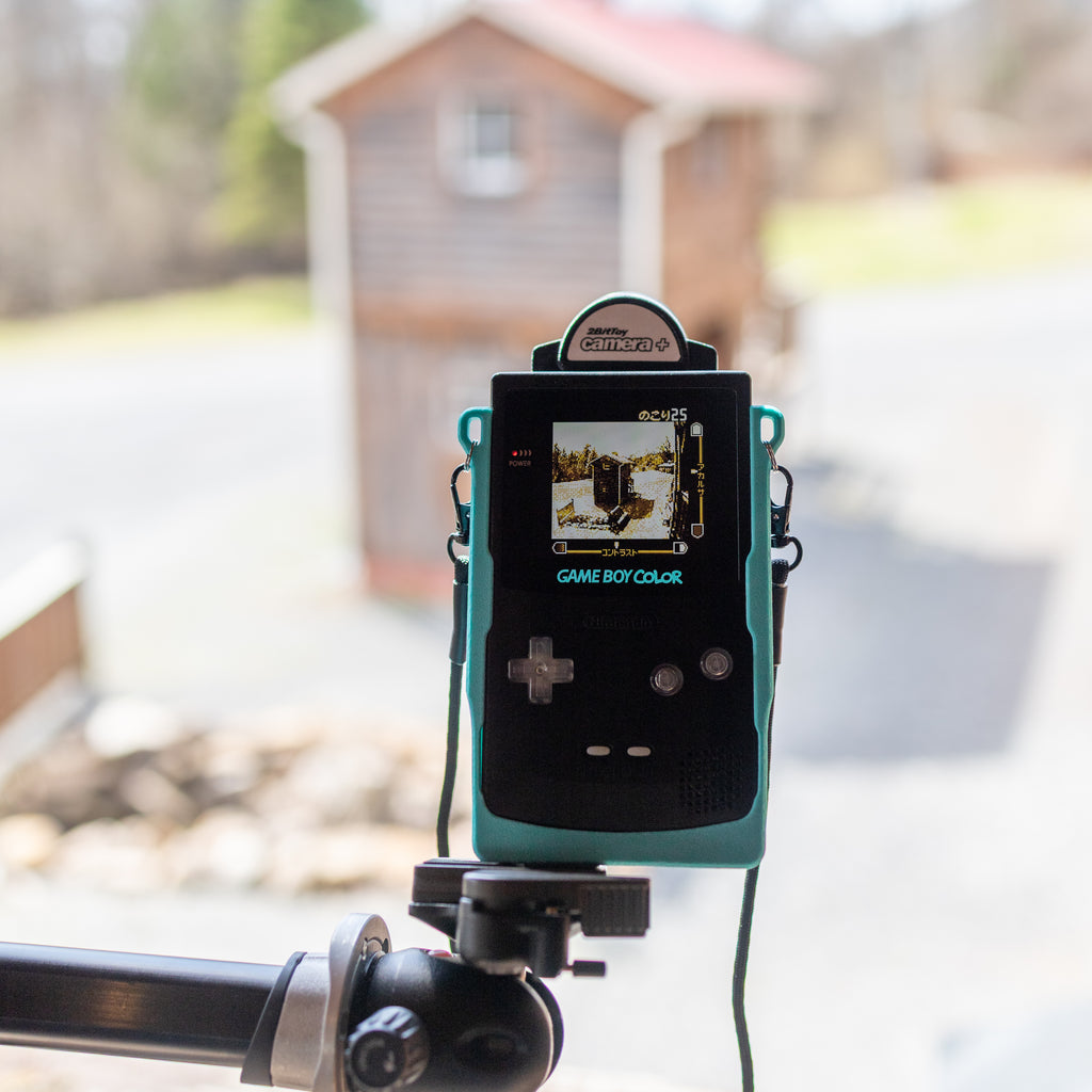 A first-person perspective looking down at a tripod-mounted black Game Boy Color in a teal 3D-printed Cradle. The screen clearly shows a pixelated 2-bit image of a small house.