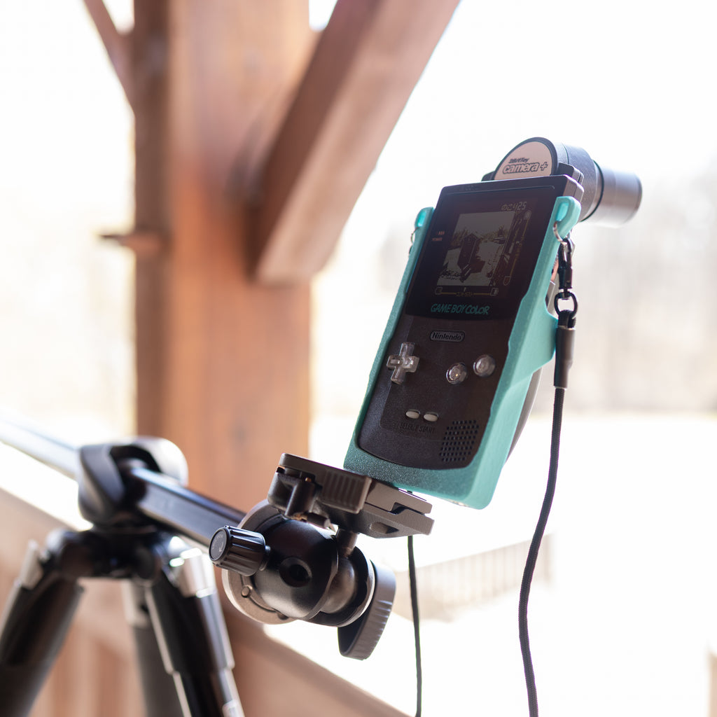 A bright, well-lit front view of a black Game Boy Color in a teal 3D-printed Cradle; the screen is visible as it captures a shot while mounted on a tripod.