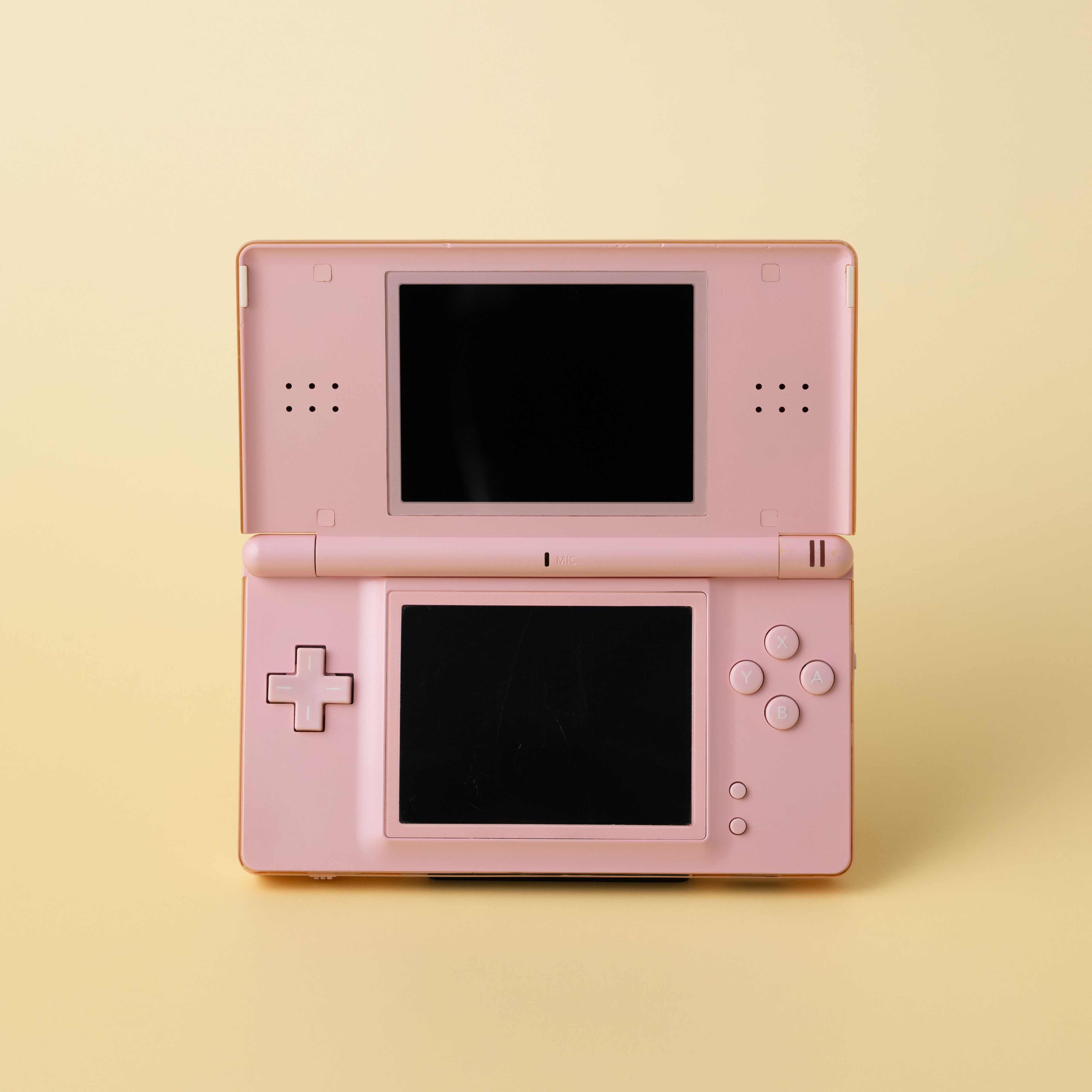 A light pink Nintendo DS Lite held by a discrete INCOG display stand on a pale yellow background.