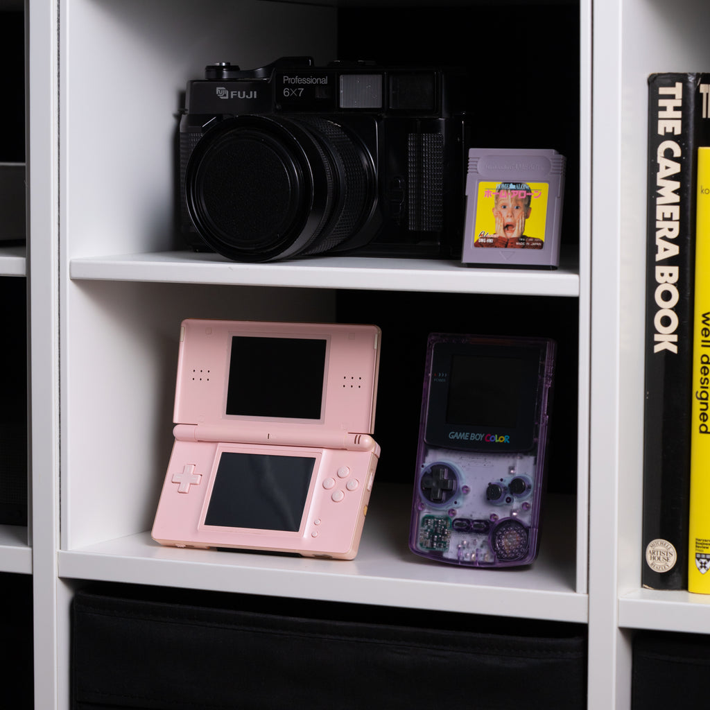 A gaming shelf featuring a pink DS Lite on an INCOG stand, an Atomic Purple GBC, a Fuji 6x7 film camera, and Home Alone for Game Boy.
