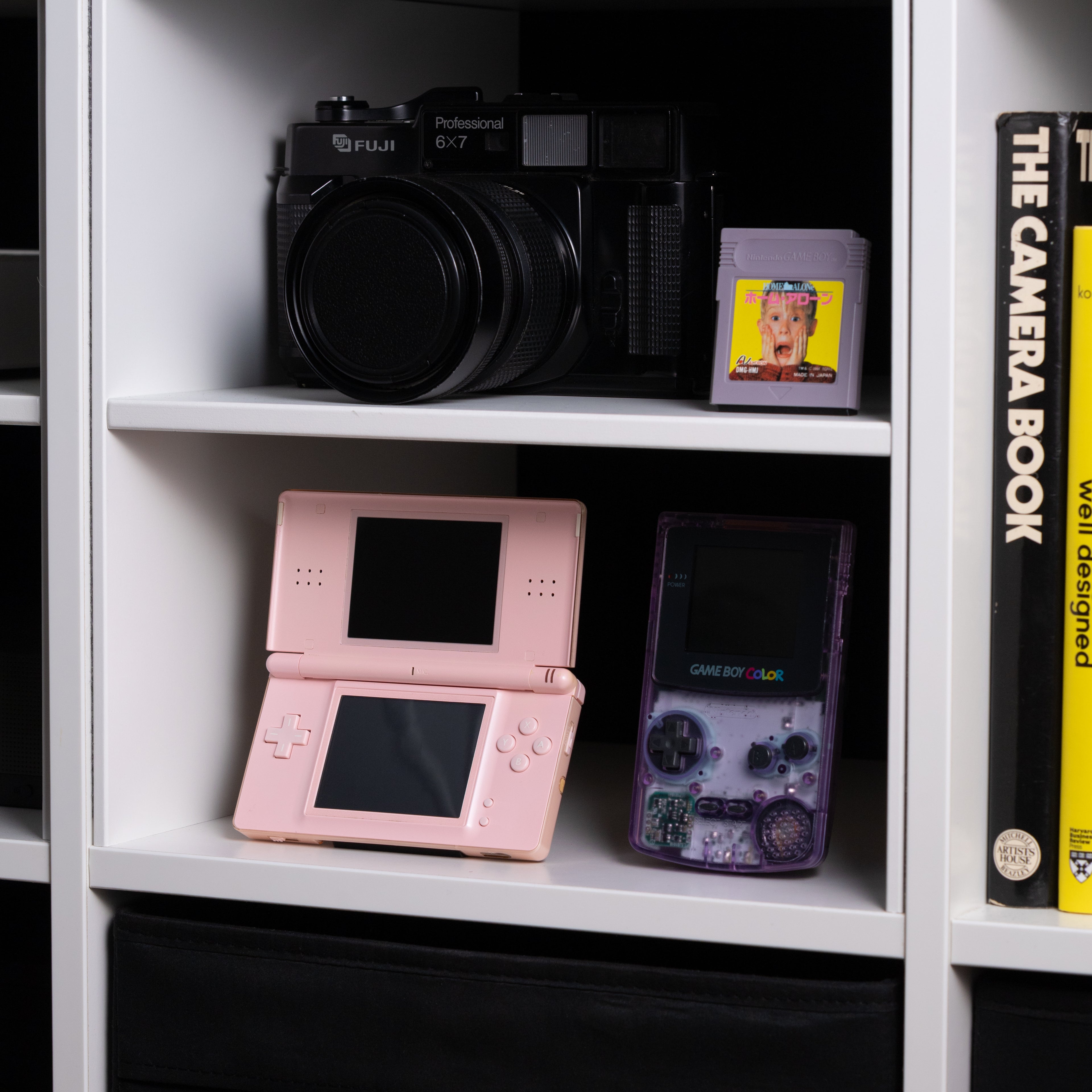 A gaming shelf featuring a pink DS Lite on an INCOG stand, an Atomic Purple GBC, a Fuji 6x7 film camera, and Home Alone for Game Boy.