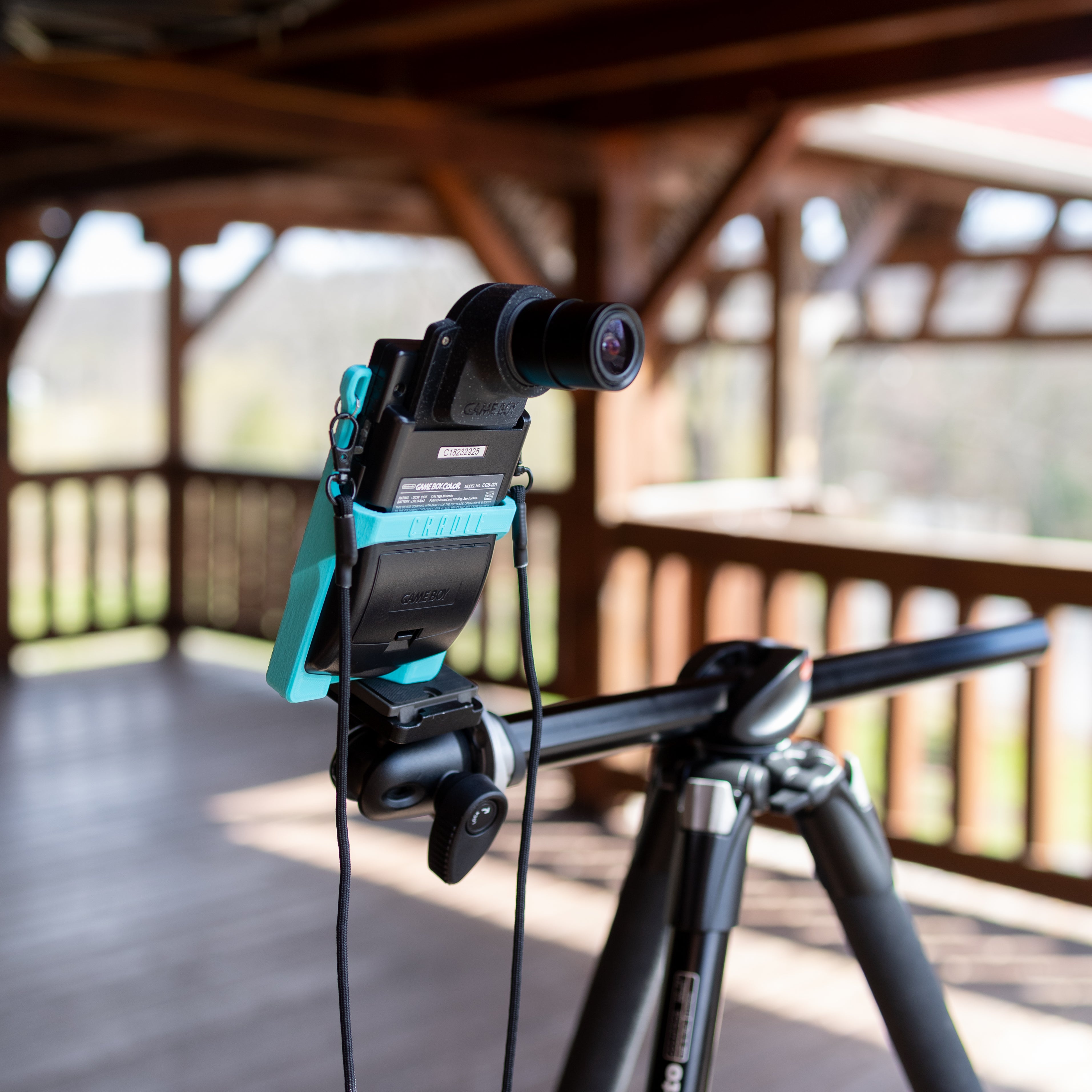 A close-up, sharp focus on a teal 3D-printed Cradle holding a black Game Boy Color on a tripod, with the wooden slats of a balcony blurred in the background.