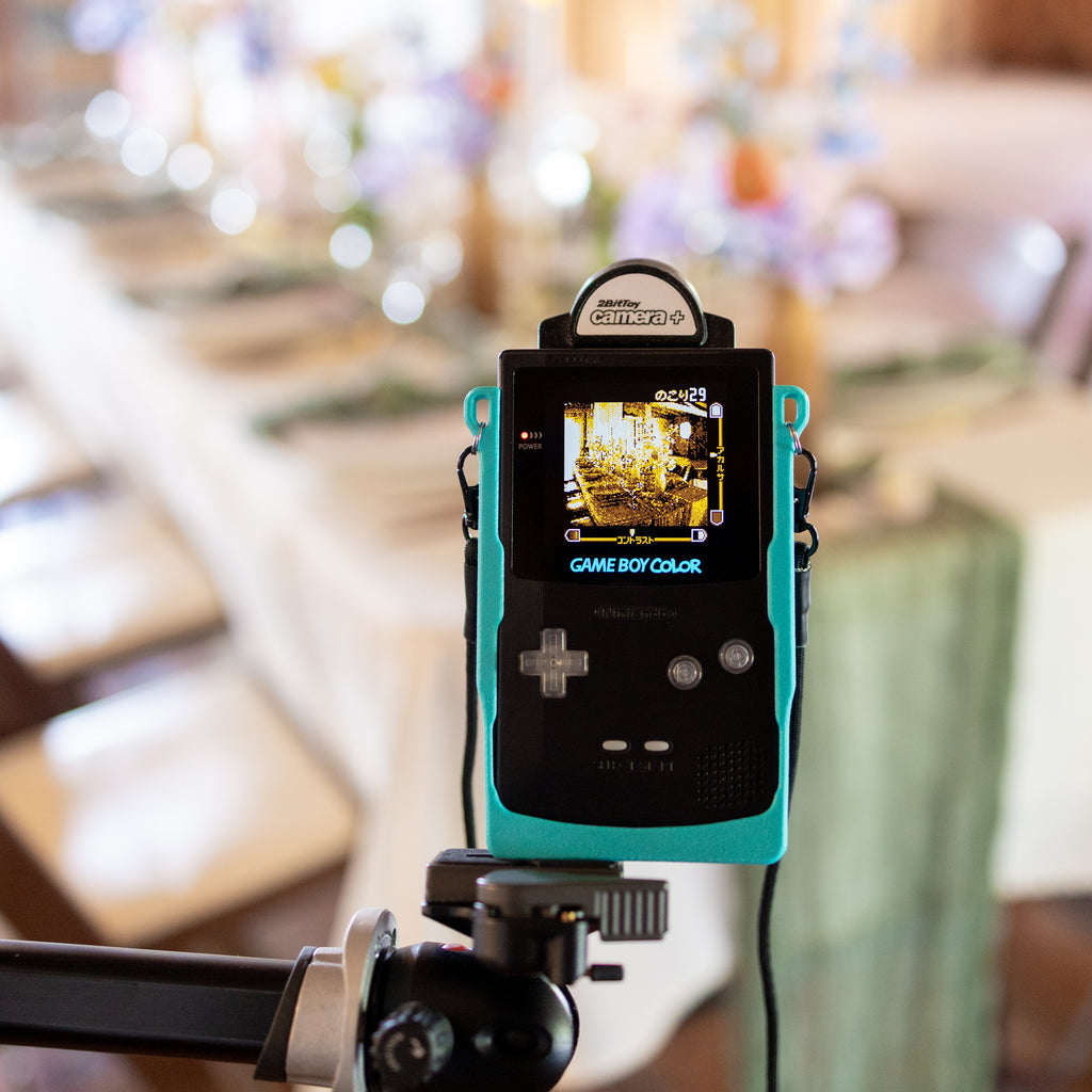 A black Game Boy Color secured in a teal 3D-printed Cradle, mounted on a tripod to photograph an elegant wedding reception table centerpiece