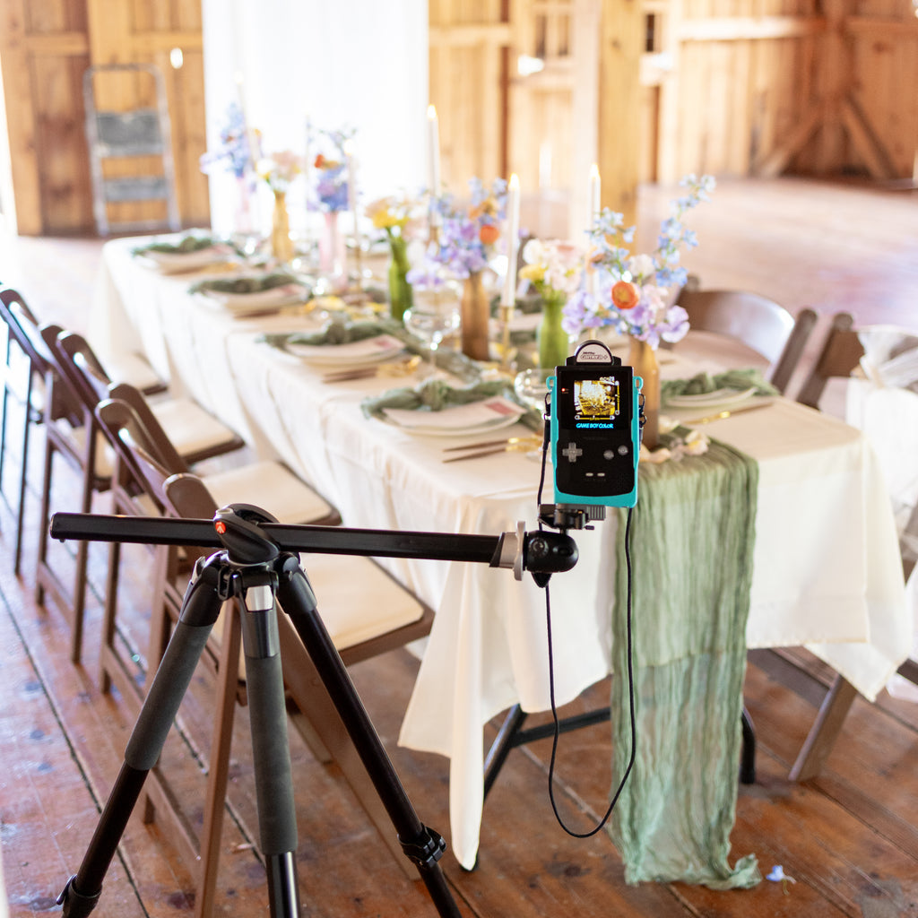 A wide shot of a black Game Boy Color and teal 3D-printed Cradle mounted on a tripod, set up to capture a detailed wedding table decoration in a bright reception hall.