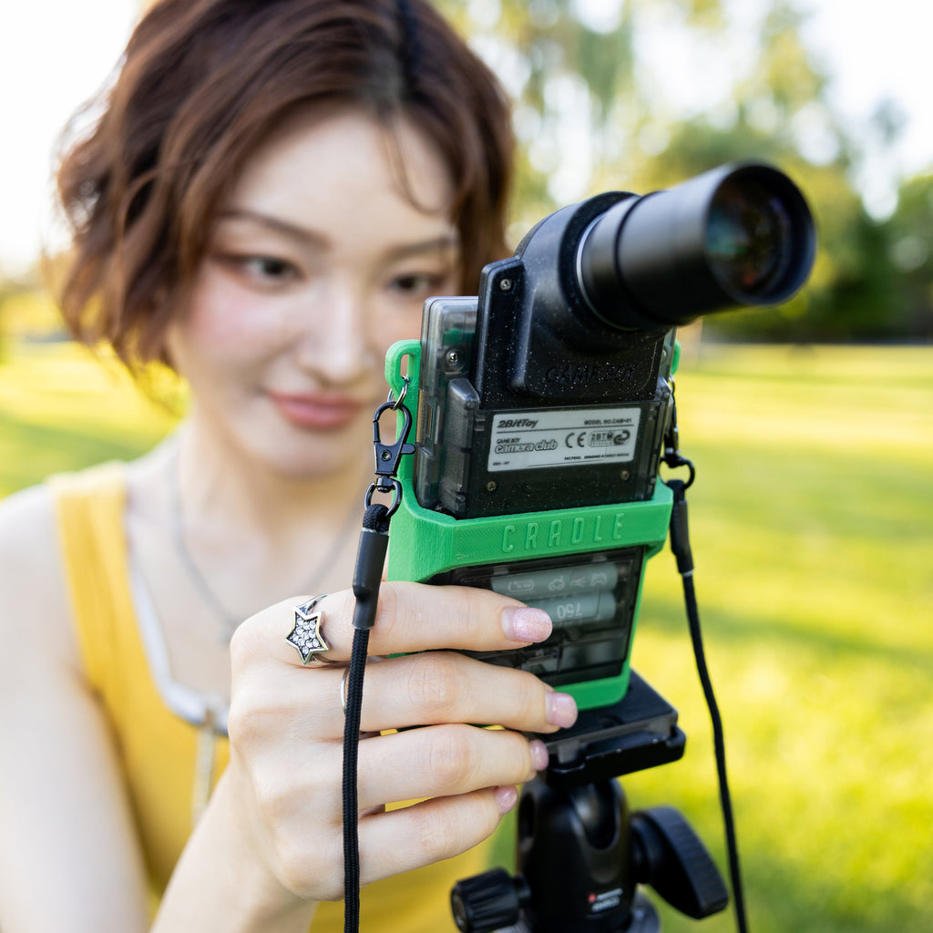 An Asian woman smiling while looking through a modified Game Boy Pocket camera at a park.